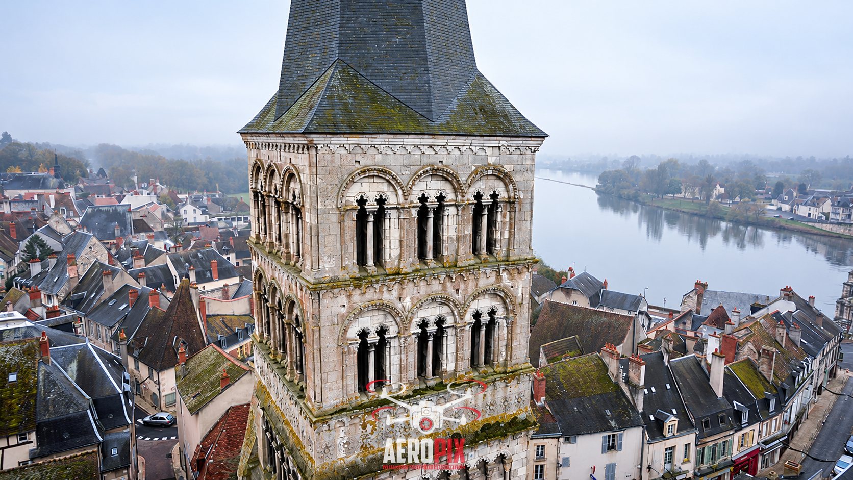 Vue aérienne par drone d'un clocher roman en pierre dominant les toits d'une petite ville et une rivière enveloppée de brume.