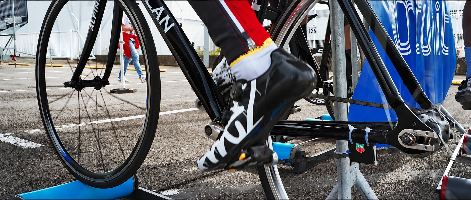 Gros plan sur le pied d'un cycliste en chaussure noire et blanche pédalant sur un vélo fixé à un home trainer bleu, sur un parking avant une course.