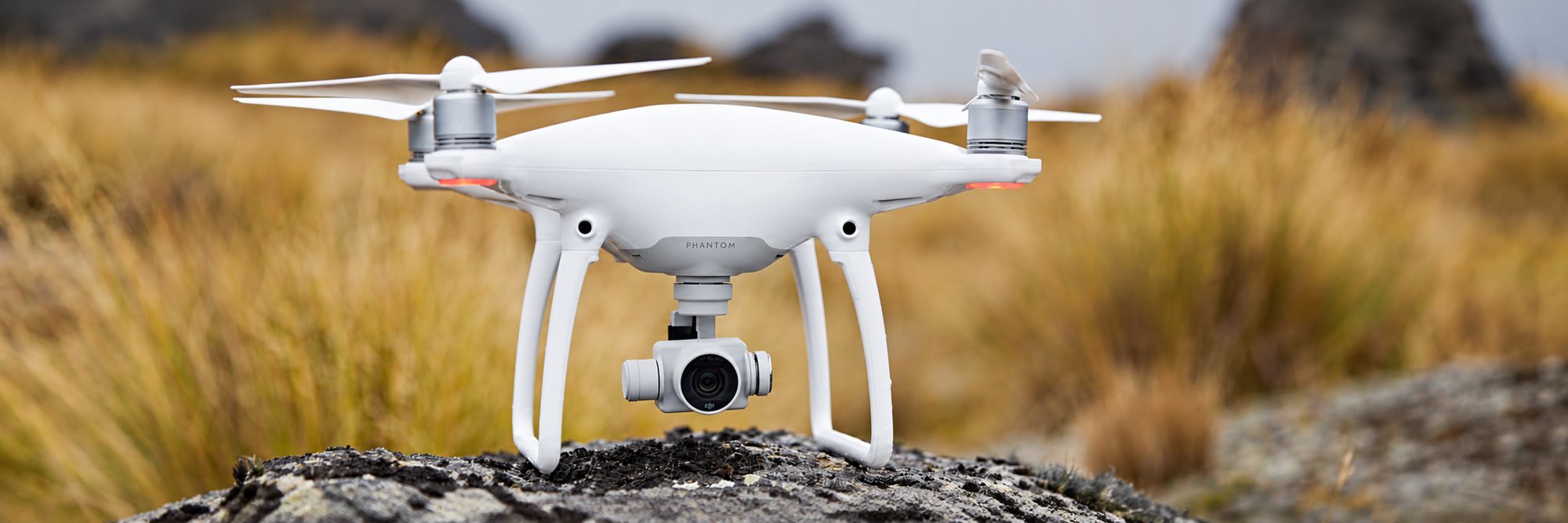 Un drone quadricoptère blanc équipé d'une caméra sur nacelle est posé sur un rocher, devant une végétation sauvage floue aux tons dorés.