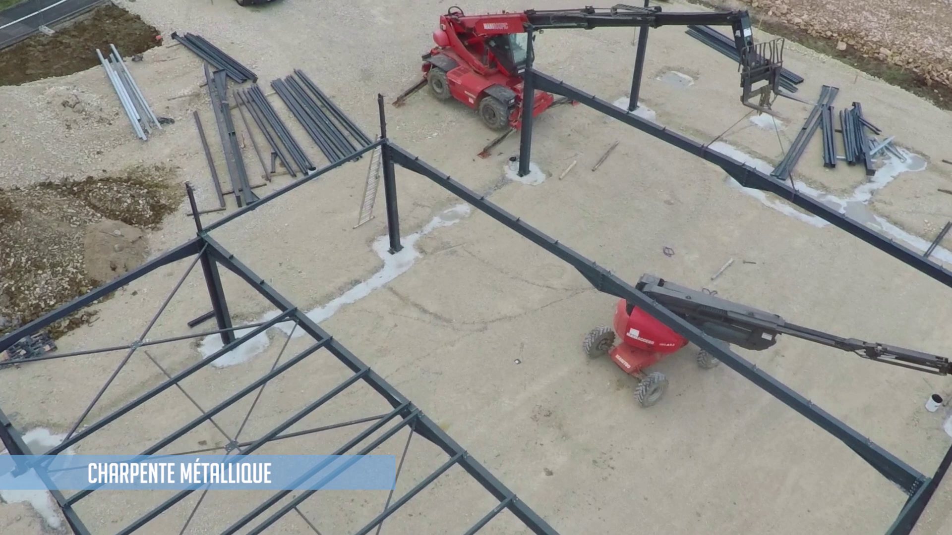 Vue aérienne d'un chantier où une charpente métallique est montée à l'aide d'un chariot télescopique rouge et d'une nacelle élévatrice, avec des poutres en acier posées au sol.