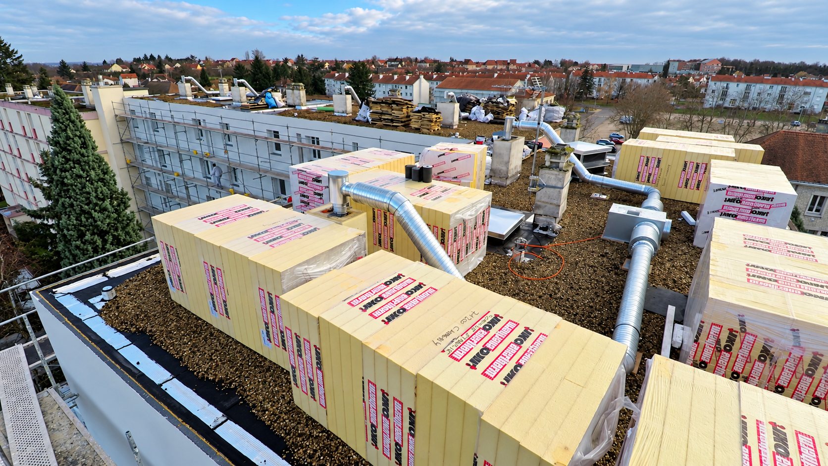 Vue aérienne d'un toit-terrasse où sont posées de nombreuses palettes de panneaux isolants jaunes, à côté d'un immeuble entouré d'échafaudages.
