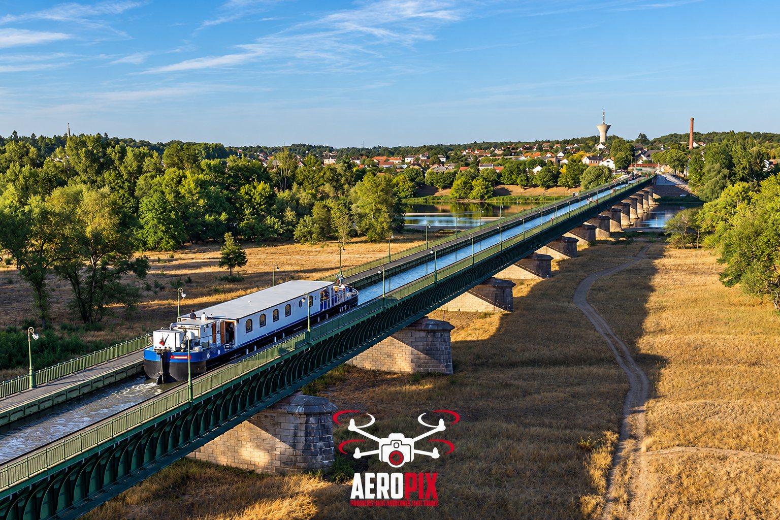 Vue aérienne par drone d'une péniche bleue et blanche naviguant sur un long pont-canal au milieu de prairies sèches, avec une ville et un château d'eau au loin.