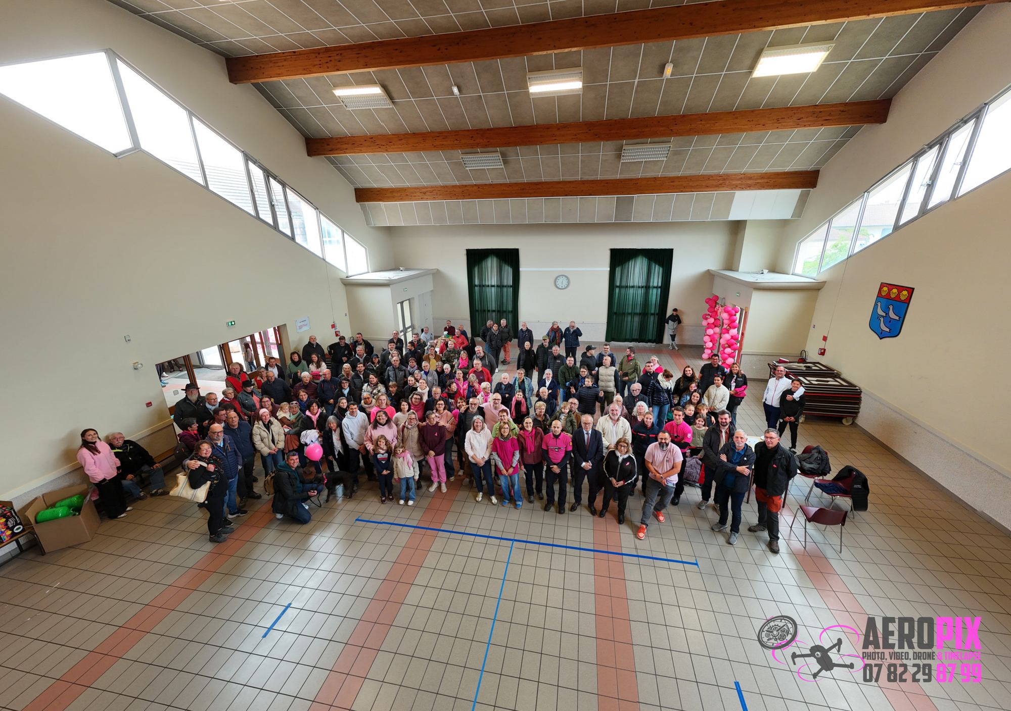 Vue en hauteur d'une grande foule rassemblée pour une photo de groupe dans une vaste salle carrelée avec des ballons roses et un blason au mur.