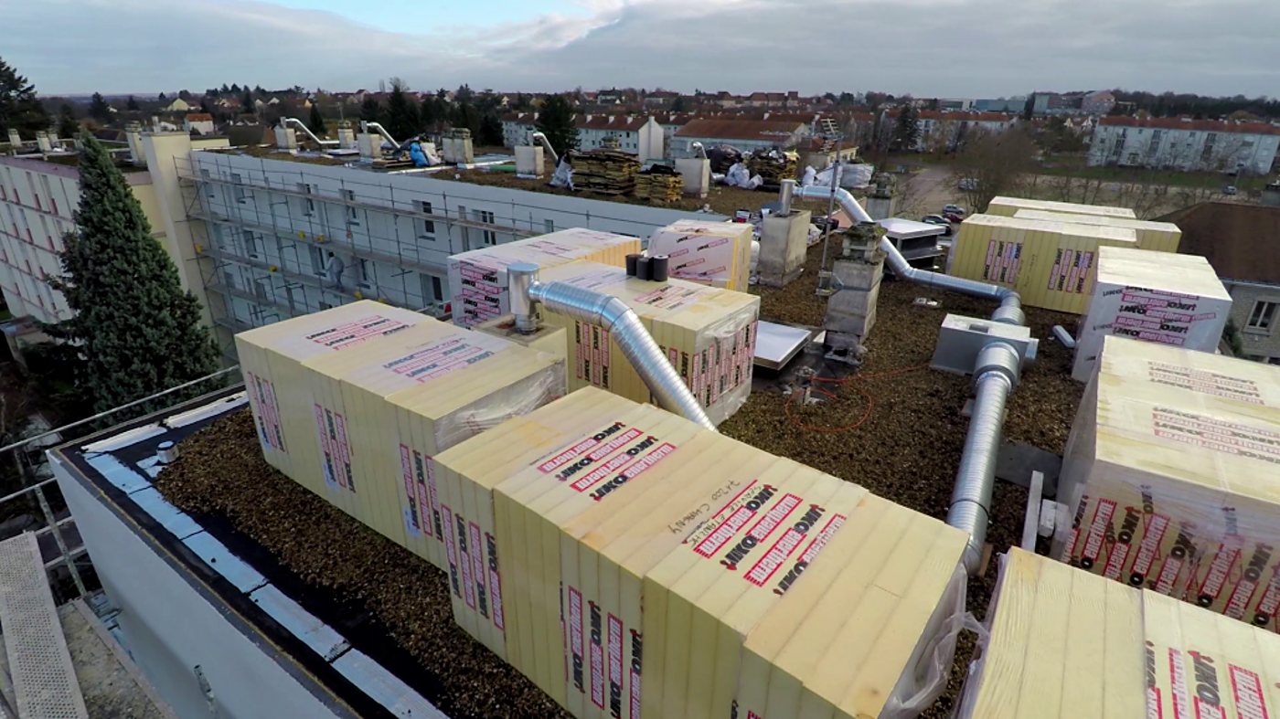 Vue aérienne d'un chantier de rénovation où des palettes de panneaux isolants jaunes sont stockées sur le toit-terrasse d'un immeuble entouré d'échafaudages.