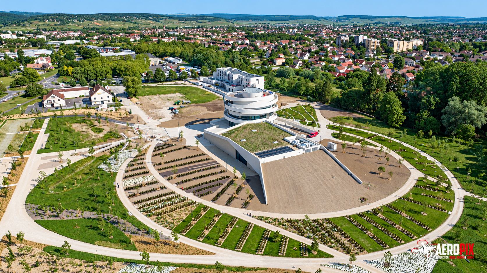 Vue aérienne par drone d'un bâtiment moderne à tour ronde blanche, entouré de jardins en terrasses circulaires et de chemins clairs, avec une ville en arrière-plan.