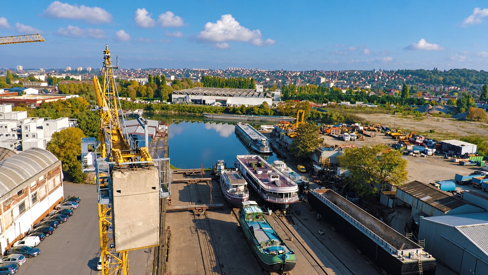 Vue aérienne d'un chantier naval au bord d'un bassin, avec une grande grue jaune, plusieurs péniches en cale sèche et des engins de chantier.