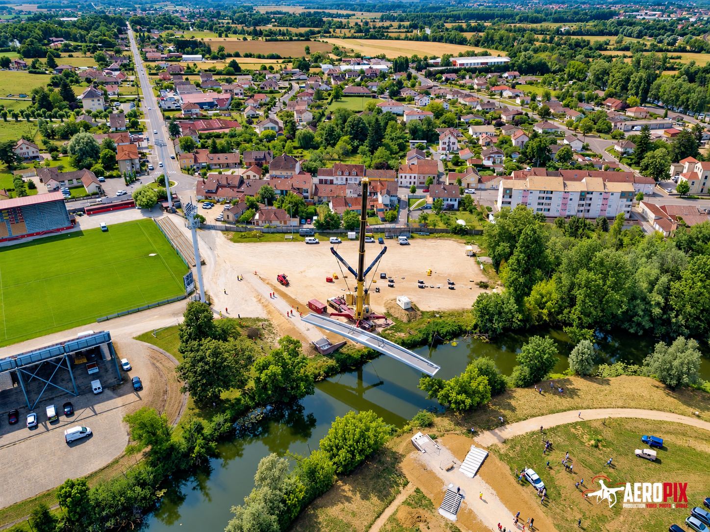 Vue aérienne par drone d'une grue installant une passerelle métallique au-dessus d'une rivière, près d'un stade et d'un quartier résidentiel.