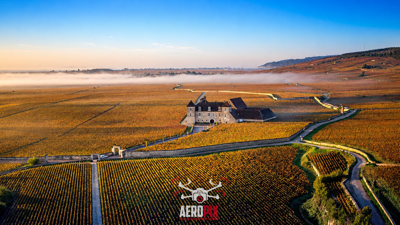 Photo prise par drone d'un château au milieu de vignobles aux couleurs d'automne, avec de la brume au loin et le logo Aeropix au premier plan.