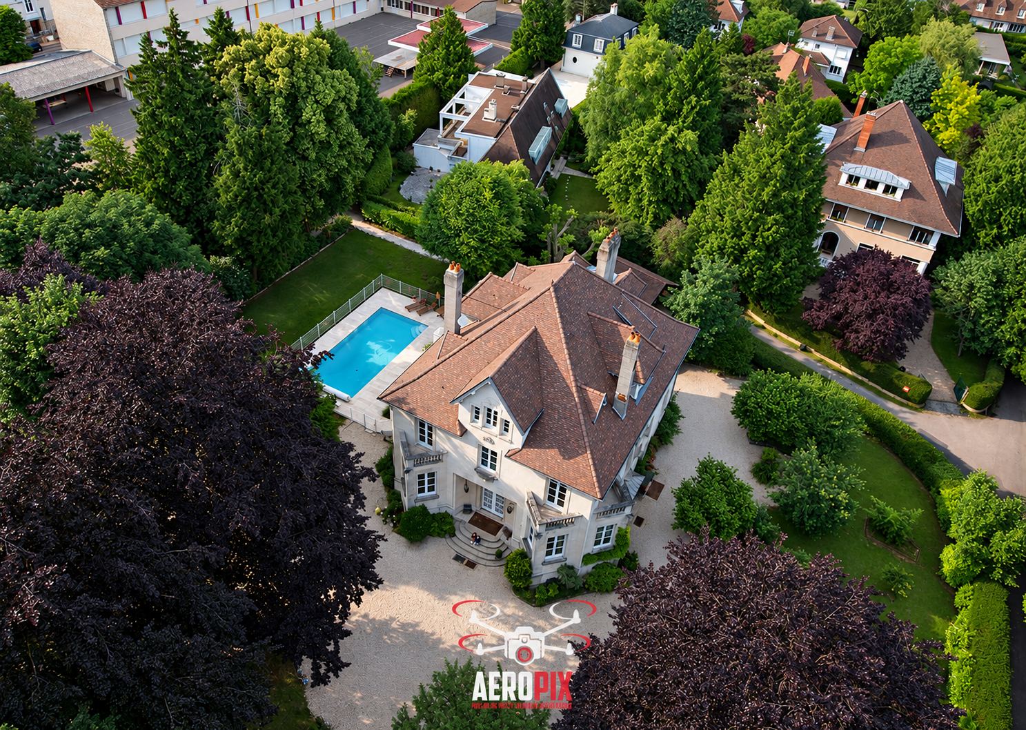 Photo prise par drone d'une grande maison bourgeoise à toit de tuiles avec piscine, entourée d'arbres et d'une allée de gravier, avec le logo Aeropix en bas.