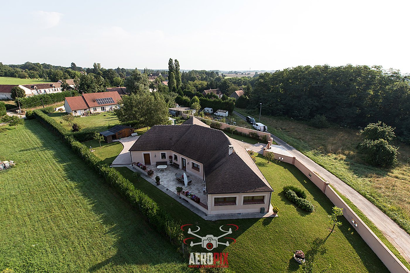 Photo prise par drone d'une maison de plain-pied à toit brun, entourée d'une pelouse, de haies et d'un mur rose, en pleine campagne.