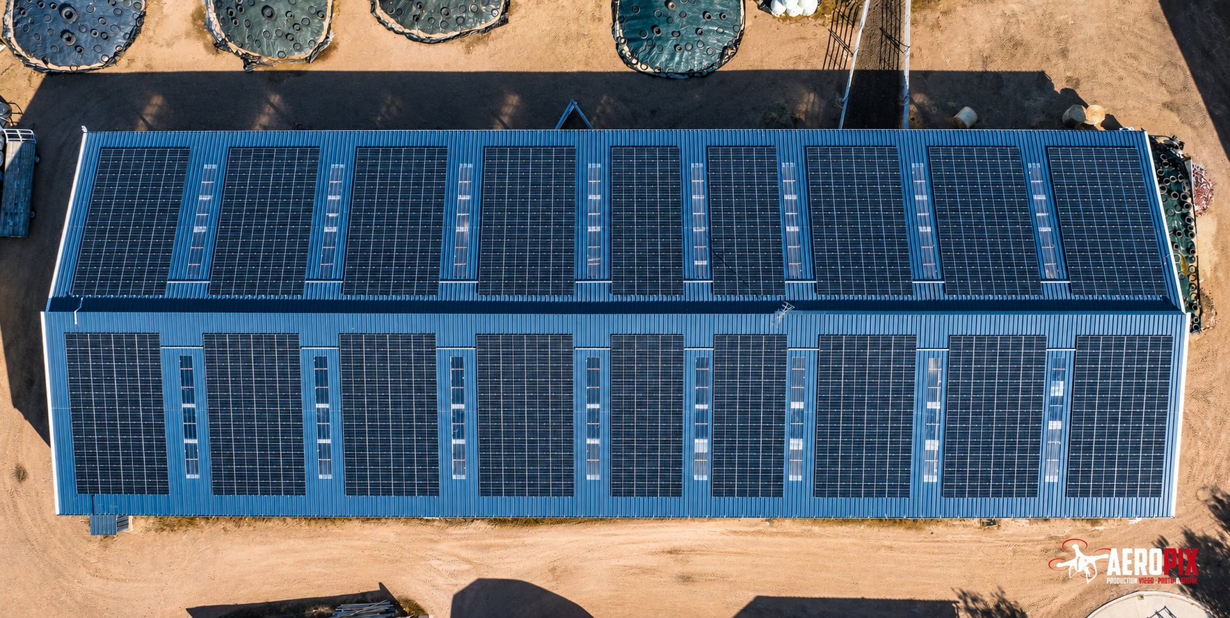 Vue de drone d'un long bâtiment au toit bleu entièrement équipé de panneaux photovoltaïques, entouré de terre sèche.