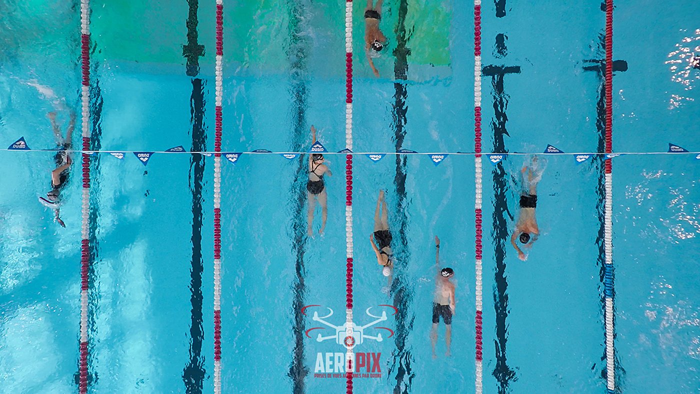 Photo prise par drone au-dessus d'une piscine, montrant plusieurs nageurs dans des couloirs séparés par des lignes d'eau rouges et blanches.