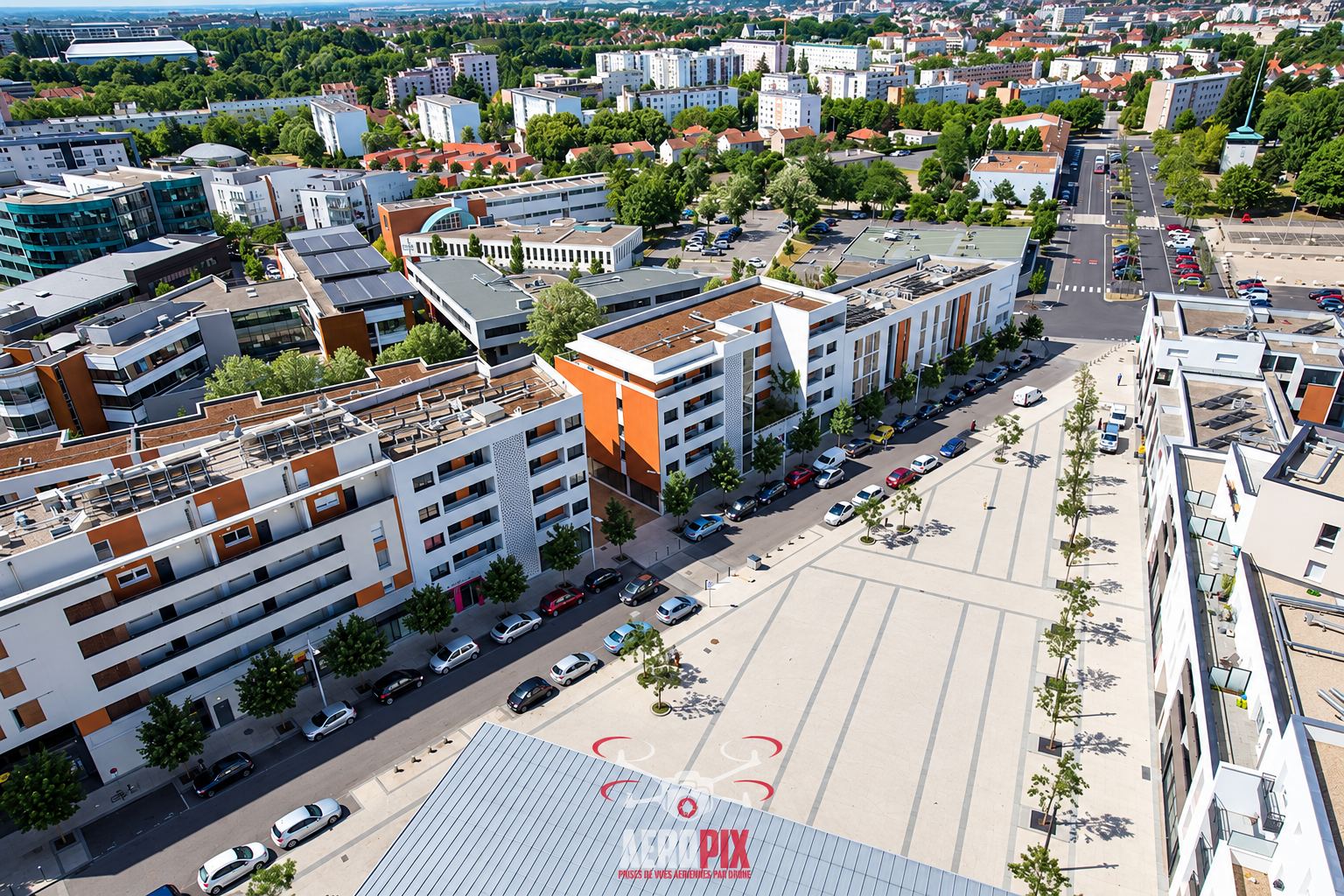 Photo prise par drone d'un quartier urbain moderne avec immeubles blancs et orange, une grande place pavée plantée de jeunes arbres et des voitures garées le long de la rue.