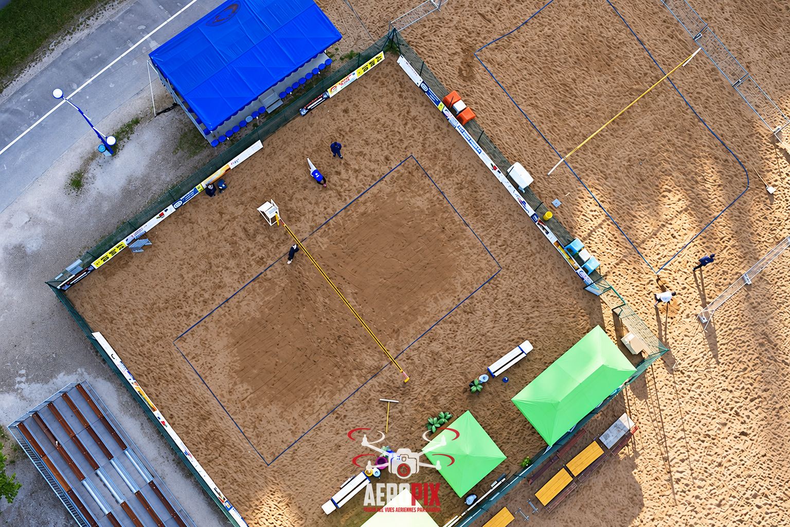 Photo prise par drone d'un terrain de beach-volley en sable avec un filet, des joueurs, des banderoles publicitaires et des tentes bleue et verte.